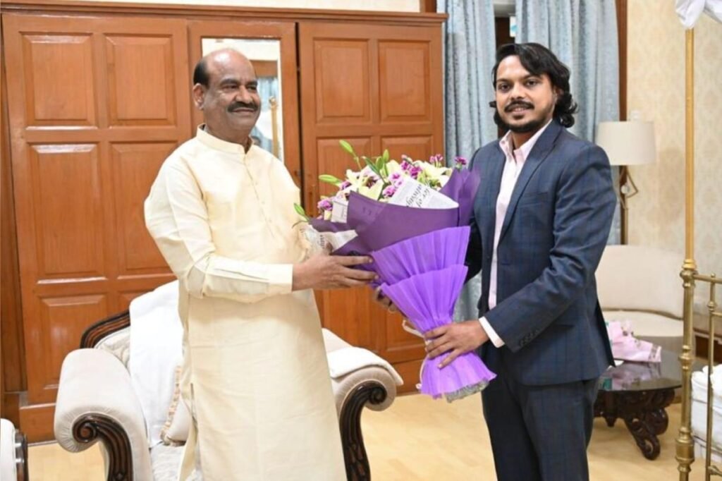 A photograph of Yash Kataria and Om Birla seated at a wooden table during a formal meeting, with the Lok Sabha complex in the background.